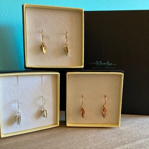 May include: Three small boxes with open lids, each containing a pair of delicate gold, silver, and rose gold leaf earrings. The boxes are lined with white velvet and have black exteriors. The boxes are arranged in a row on a wooden surface. The text "silverbee" is printed on the black box.