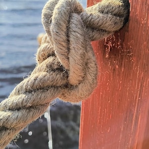 May include: A thick, natural-coloured fibre rope is tied in a secure knot through a circular opening in a vertical, weathered orange-red wooden post. The rope shows a textured, twisted construction, and the post has a visible wood grain finish.