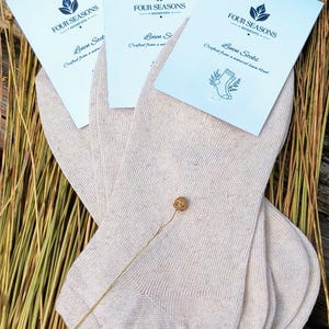 Pode incluir: Três pares de meias curtas bege claro com etiquetas de papel branco marcadas como Four Seasons Moments Linen Socks. As meias têm uma textura de malha fina e são exibidas com um pequeno caule de planta seca sobre o tecido.