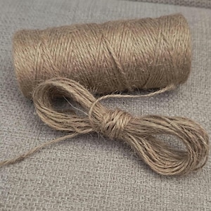 May include: A cylindrical spool of natural light brown jute twine featuring a textured, fibrous surface. A loose length of the same brown twine is coiled into a small, oval-shaped loop positioned directly in front of the main spool.