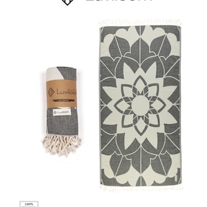 May include: A charcoal gray and off-white Turkish towel featuring a large symmetrical floral mandala pattern and tasseled ends. One towel is displayed flat, while another is rolled and secured with a branded paper band labeled Luviloom.