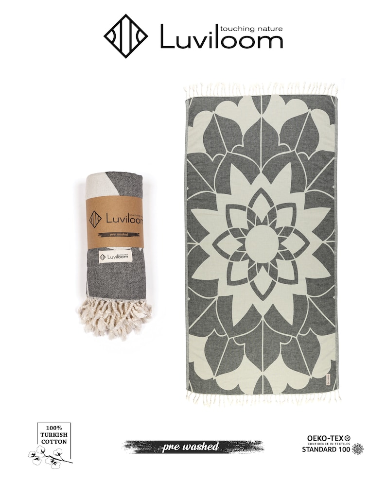 May include: A charcoal gray and off-white Turkish towel featuring a large symmetrical floral mandala pattern and tasseled ends. One towel is displayed flat, while another is rolled and secured with a branded paper band labeled Luviloom.