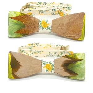 Può includere: Due papillon in legno con motivo floreale sul cinturino e sul cinturino. I papillon hanno una forma rettangolare con finitura a venatura del legno marrone chiaro e accenti in resina verde e gialla alle estremità. I cinturini sono color crema con motivi floreali gialli.