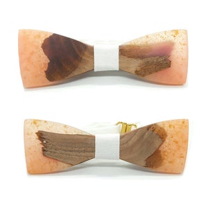 Può includere: Due papillon in legno con un nastro bianco. I papillon sono in legno marrone chiaro con un'intarsio in resina rosa.