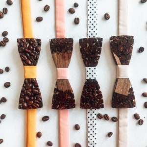 Può includere: Quattro papillon in legno con chicchi di caffè incorporati nel legno. I papillon sono esposti su nastri di diversi colori: arancione, rosa, bianco con pois neri e beige.