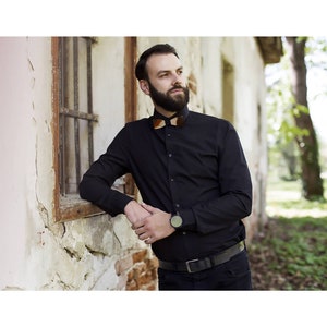 Può includere: Un uomo indossa una camicia nera abbottonata con un papillon di legno. È in piedi accanto a un muro di mattoni e guarda a destra. Indossa un orologio nero al polso sinistro.