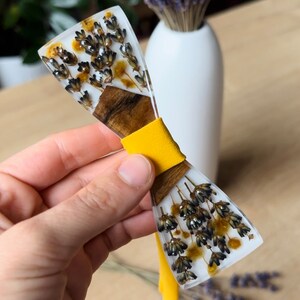 Può includere: Un papillon in resina bianca con fiori di lavanda e gialli secchi incorporati nella resina. Il papillon ha un cinturino in tessuto giallo.