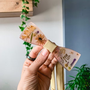 Può includere: Un papillon rosa chiaro con fiori pressati racchiusi in resina. Il papillon ha un cinturino in raso giallo.