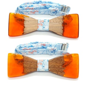 Può includere: Due papillon in legno con dettagli in resina arancione. I papillon hanno un nastro in tessuto con motivo floreale blu chiaro.