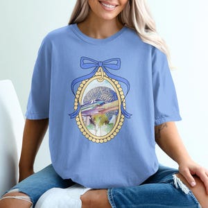 May include: A light blue t-shirt featuring a framed illustration of a theme park with a large geodesic sphere and colorful accents. The frame is adorned with a blue bow and a pearl-like border. The shirt is a solid light blue color.