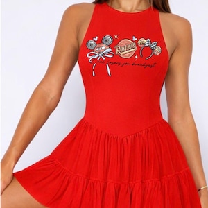 May include: A vibrant red dress with a fitted bodice and a flared, tiered skirt. The sleeveless dress features a graphic print of a cartoon car, a logo, and text. The overall design is playful and eye-catching.