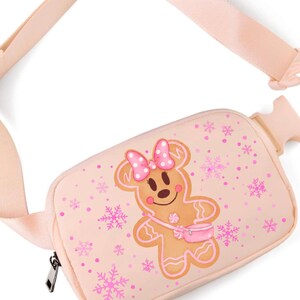 May include: A light pink fanny pack with a gingerbread woman design. The gingerbread woman has a pink bow and purse. Pink snowflakes and dots are scattered across the bag. The adjustable strap and zipper are also light pink.