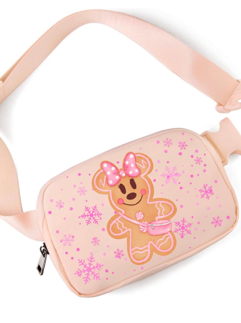 May include: A light pink fanny pack with a gingerbread woman design. The gingerbread woman has a pink bow and purse. Pink snowflakes and dots are scattered across the bag. The adjustable strap and zipper are also light pink.