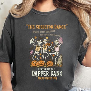 May include: Dark gray t-shirt featuring a Halloween design. The shirt displays the text "THE SKELETON DANCE" above a graphic of dancing skeletons playing instruments, pumpkins, and a tombstone. The text "FEATURING THE DAPPER DANS MAIN STREET USA" is also included.