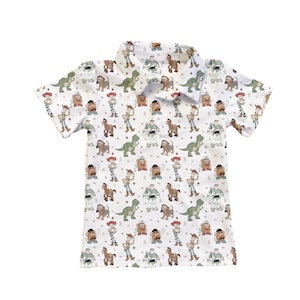 May include: White short-sleeved polo shirt with a repeating pattern of Toy Story characters. The design includes images of Woody, Buzz Lightyear, Rex, Mr. Potato Head, and other characters from the animated film. The shirt has a collared neckline.