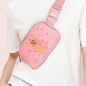 May include: A pink fanny pack with a gingerbread person graphic and snowflake accents. The bag has a zipper closure and an adjustable strap with a buckle. The gingerbread person has a pink bow and scarf.