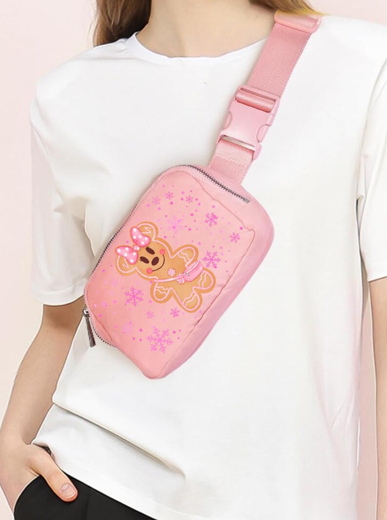 May include: A pink fanny pack with a gingerbread person graphic and snowflake accents. The bag has a zipper closure and an adjustable strap with a buckle. The gingerbread person has a pink bow and scarf.
