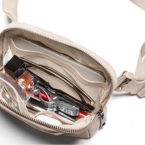 May include: A beige crossbody bag with a zip closure, revealing its contents. The bag features an adjustable strap and a plastic buckle. Inside, there are sunglasses, a watch, a small perfume bottle, and other personal items.