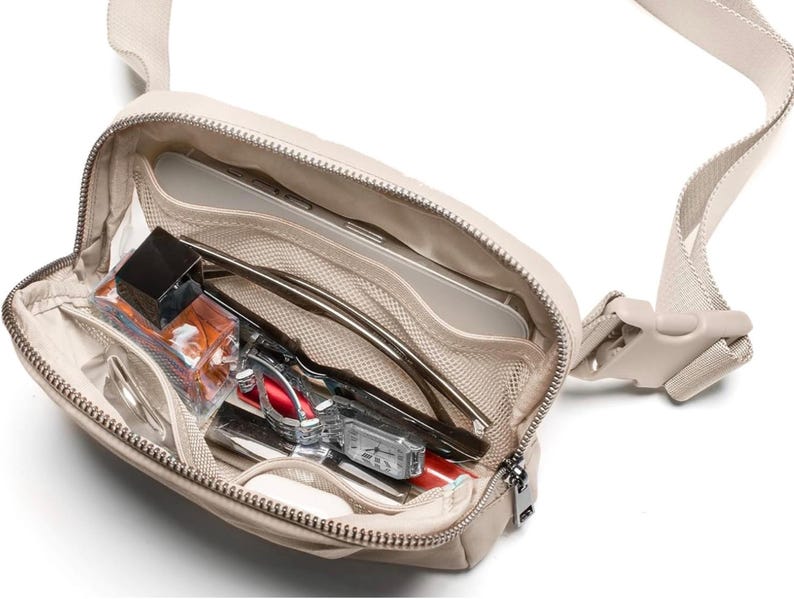 May include: A beige crossbody bag with a zip closure, revealing its contents. The bag features an adjustable strap and a plastic buckle. Inside, there are sunglasses, a watch, a small perfume bottle, and other personal items.