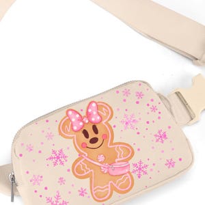 May include: Beige fanny pack with a gingerbread cookie design. The gingerbread character has a pink bow, purse, and is surrounded by pink snowflakes and dots. The strap and buckle are beige.