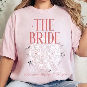 May include: A light pink t-shirt with the text "THE BRIDE" in a reddish-pink font. Below the text is a geometric design with small illustrations and the phrase "Celebrating Happily Ever After."