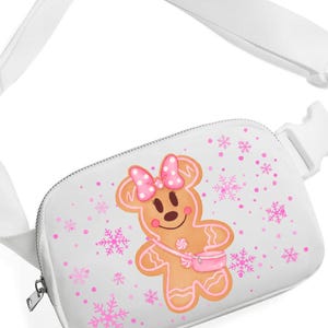 May include: White fanny pack with a gingerbread person graphic. The gingerbread person has a pink bow, a pink purse, and pink accents. Pink snowflakes and dots are scattered across the white background. The strap and buckle are white.