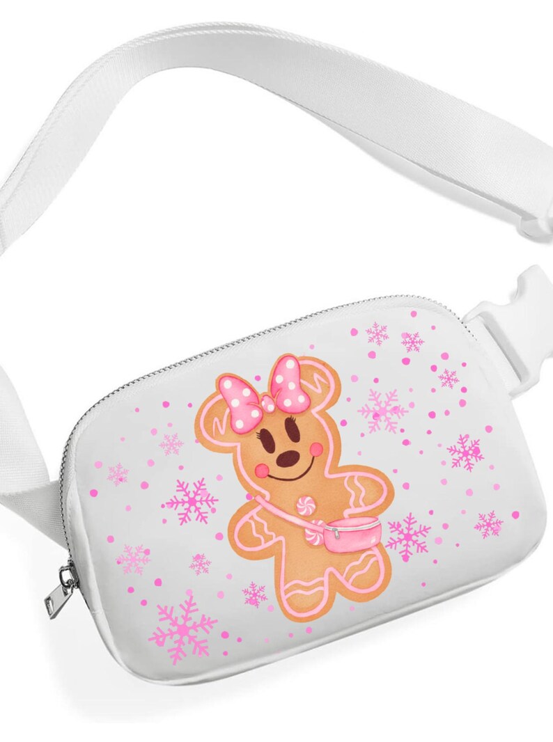May include: White fanny pack with a gingerbread person graphic. The gingerbread person has a pink bow, a pink purse, and pink accents. Pink snowflakes and dots are scattered across the white background. The strap and buckle are white.