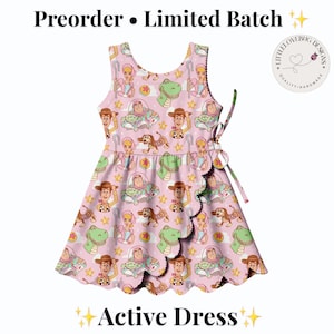 May include: A sleeveless pink active dress featuring a repeating pattern of cartoon characters including a cowboy, a space ranger, a dinosaur, and a dog. The dress has a wrap-style front with a scalloped hem and a side tie closure. Text reads Preorder Limited Batch.