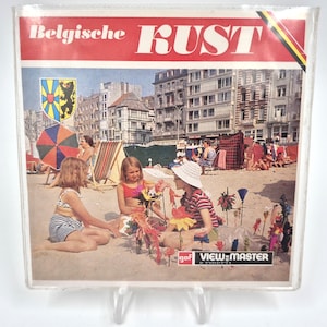 Pode incluir: Um carretel View-Master vintage com uma cena de praia com crianças brincando na areia. O carretel tem o texto "Belgische Kust" em vermelho, com um cenário de cidade costeira e um guarda-sol colorido. A imagem está dentro de um suporte de plástico transparente.