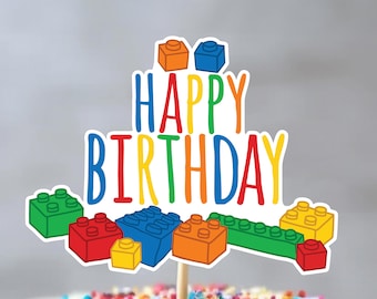 Op de afbeelding: Een kleurrijke taarttopper met de woorden "Happy Birthday" in regenboogkleuren en een ontwerp van kleurrijke Lego-blokken.