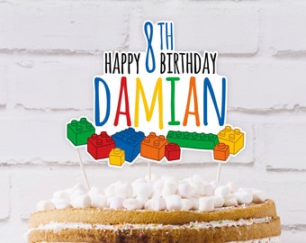 May include: A birthday cake topped with a colorful cake topper. The topper features the text "Happy 8th Birthday Damian" in blue, red, yellow, and green, with Lego-style block graphics. The cake is decorated with marshmallows.