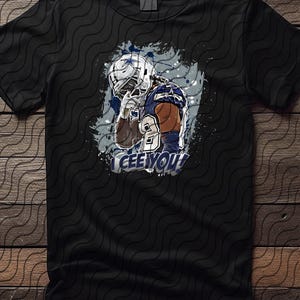 May include: Black t-shirt featuring a graphic of a football player in a helmet and uniform. The player is depicted in a thoughtful pose, with the text "CEE YOU!" above the image. The design includes blue and white accents.