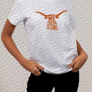 May include: White t-shirt with a brown graphic of a longhorn steer head with the text "We Dem Horns" printed on it.