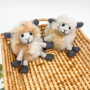 May include: Two plush sheep toys with fluffy, light brown and white wool, gray ears, and gray feet, resting in a woven basket. The sheep have black eyes and stitched facial features. The basket is made of woven, light brown material.