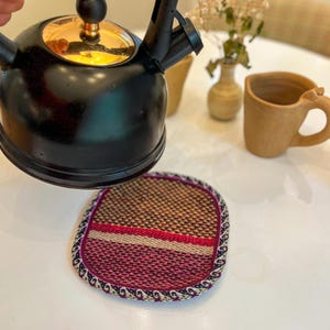 May include: A black tea kettle with a gold-colored lid is positioned above a woven coaster. The coaster has a brown, red, and white pattern with a decorative border. A ceramic mug and vase are in the background.