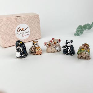 Könnte beinhalten: Satz mit sechs kleinen Keramik-Tierfiguren, darunter ein Pinguin, eine Eule, ein Koala, ein Panda und eine Schildkröte. Die Figuren sind in realistischen Farben bemalt und haben einen verspielten, handgefertigten Look. Die Figuren sind in einer rosa Box mit dem Text "artesana love" verpackt.