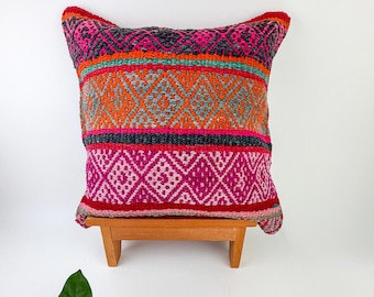 May include: A square throw pillow featuring a woven geometric pattern in vibrant shades of hot pink, bright orange, teal, and dark gray. The textile design consists of horizontal bands with diamond motifs and textured stripes, resting on a small wooden stand.