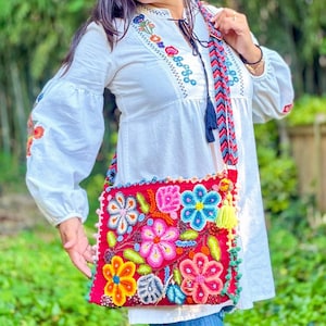 May include: A red crossbody bag with colorful floral embroidery. The bag features a braided strap with tassels and is adorned with various flower designs in shades of pink, blue, yellow, and green. The bag is trimmed with a green border.