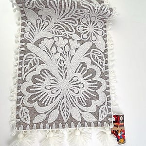 May include: A gray and white floral embroidered table runner with a repeating pattern of flowers and leaves. The runner has a white fringe trim and white tassels at the ends. The runner is approximately 60 inches long.