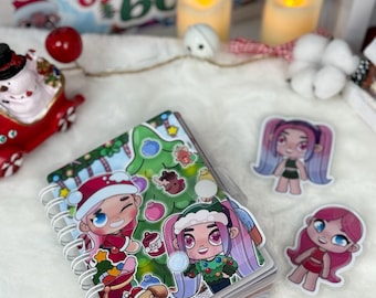 May include: A spiral-bound notebook featuring a colorful Christmas-themed illustration of cartoon characters, a decorated tree, and a puppy. Two separate die-cut stickers of cartoon characters with pink and purple hair are placed nearby.