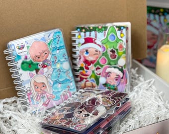 May include: A collection of spiral-bound notebooks with colorful Christmas-themed illustrations. The notebooks feature cartoon characters and festive designs, including Christmas trees and Santa hats. They are displayed in a white box with paper filler.