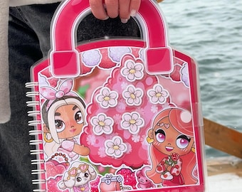 May include: A pink, handbag-shaped notebook with a clear plastic cover and a handle. The cover features illustrations of two cartoon girls, flowers, and a small dog. The notebook is held by a person wearing a white jacket.