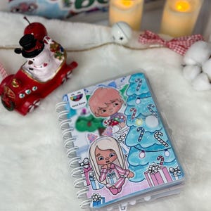 May include: A spiral-bound notebook with a clear plastic cover featuring cartoon illustrations of a Christmas tree, gifts, and characters. The cover has a white spiral binding. A red snowman figurine and candles are in the background.