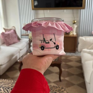 May include: A clear glass jar with a pink gingham fabric cover featuring an embroidered pink golf cart design. The jar has a ruffled pink fabric trim around the top. The jar is held in front of a blurred background.