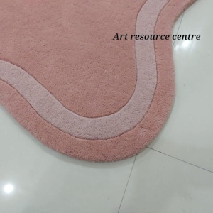 Puede incluir: Primer plano de una alfombra de ba&ntilde;o rosa y blanca con un borde ondulado. La alfombra est&aacute; hecha de un material suave y esponjoso.