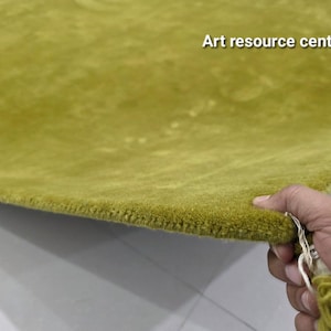 May include: A solid olive green rug with a thick, tufted pile texture. The rug features a rounded corner and a small attached tassel of matching green yarn. The text Art resource centre is printed in white in the upper right corner of the rug.