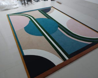 May include: Abstract geometric rug with a teal, green, pink, white, and black design on a beige background. The rug has a brown and gold border.