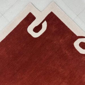 Puede incluir: Una alfombra roja con un borde blanco y un dise&ntilde;o abstracto blanco. El dise&ntilde;o presenta una forma grande y curva que se asemeja a una letra "C".