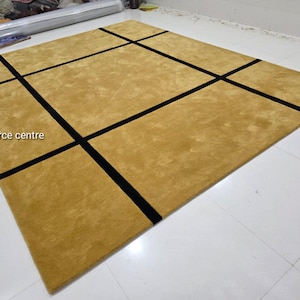 May include: A rectangular mustard-colored rug with a geometric black design. The rug features thick black lines that intersect to create a grid-like pattern. The text "Art resource centre" is visible in the lower left corner.