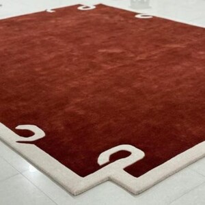 Puede incluir: Una alfombra roja con un borde blanco que presenta un dise&ntilde;o geom&eacute;trico. La alfombra tiene una textura suave y mullida.